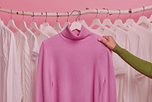 Mano que coge un jersey rosa de una percha con un fondo de camisas y camisetas colgas de sus perchas de colores blancos